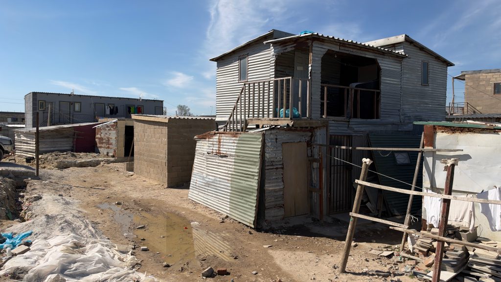 A two-storey corrugated iron dwelling beside brick and metal homes in Siyahlala informal settlement, Dunoon, with standing water on the unpaved ground.